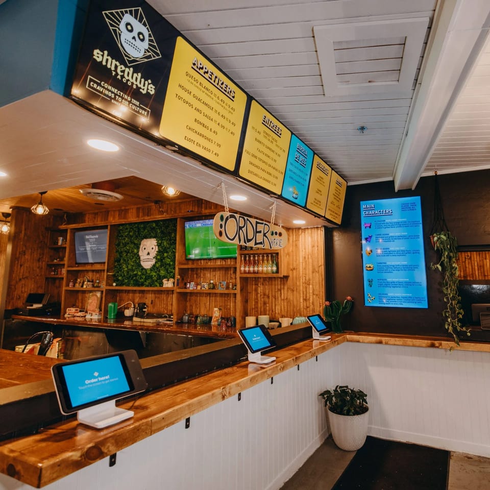 Shreddy's Tacos interior with menu boards, order kiosks, and a wooden counter with a skull logo.